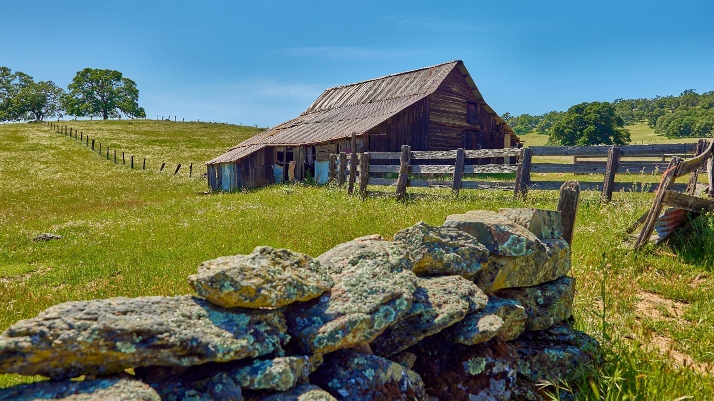A weathered barn at El Dorado Ranch behind a foreground of dry-stack rock wall and green meadow.